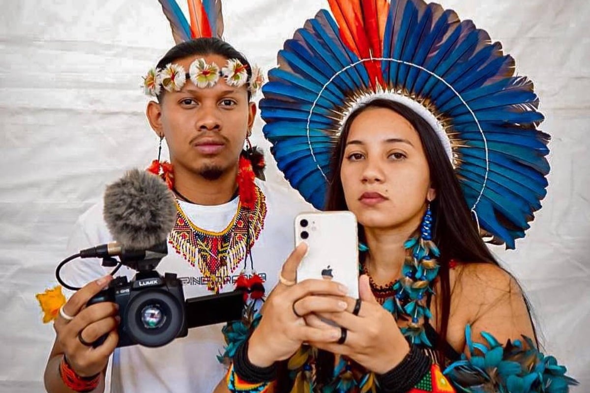 Foto de Tukumã Pataxó usando cocar, colares, blusa branca e segurando câmera, além de Samela Sateré Mawé usando cocar azul, brincos e roupa azul, segurando celular