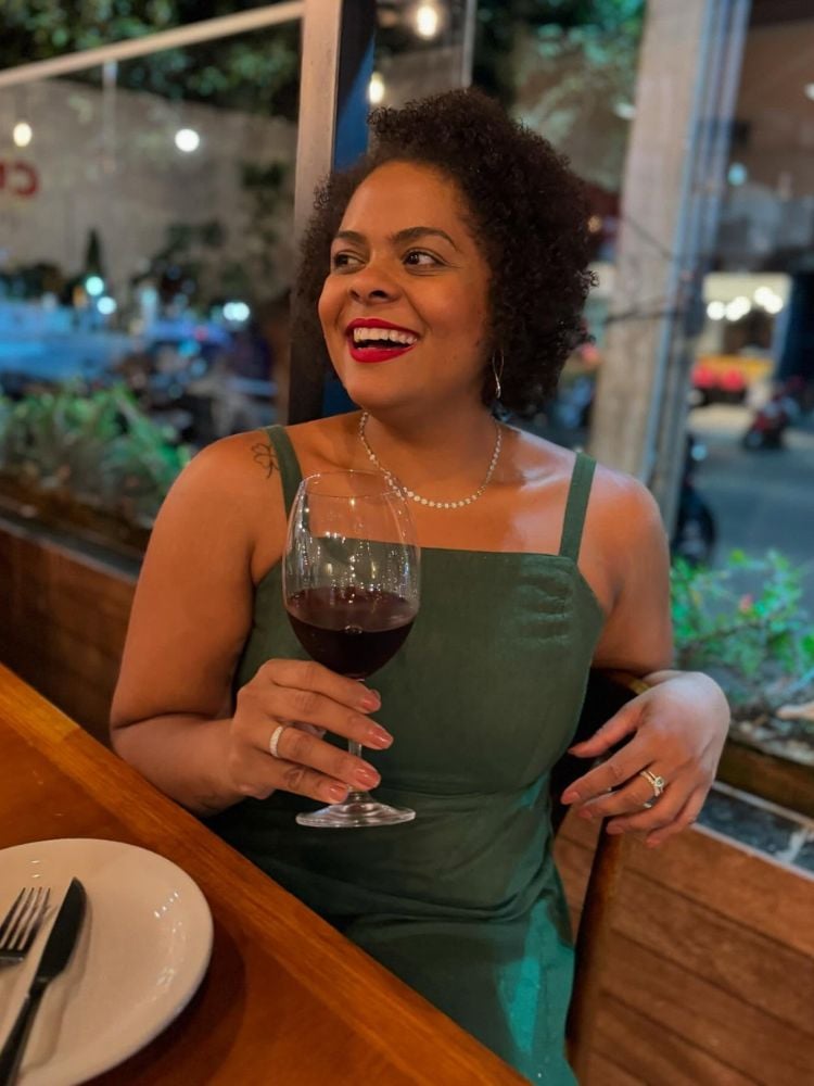 Tairine Ceuta usando vestido verde de alças finas, sentada à mesa com taça de vinho na mão