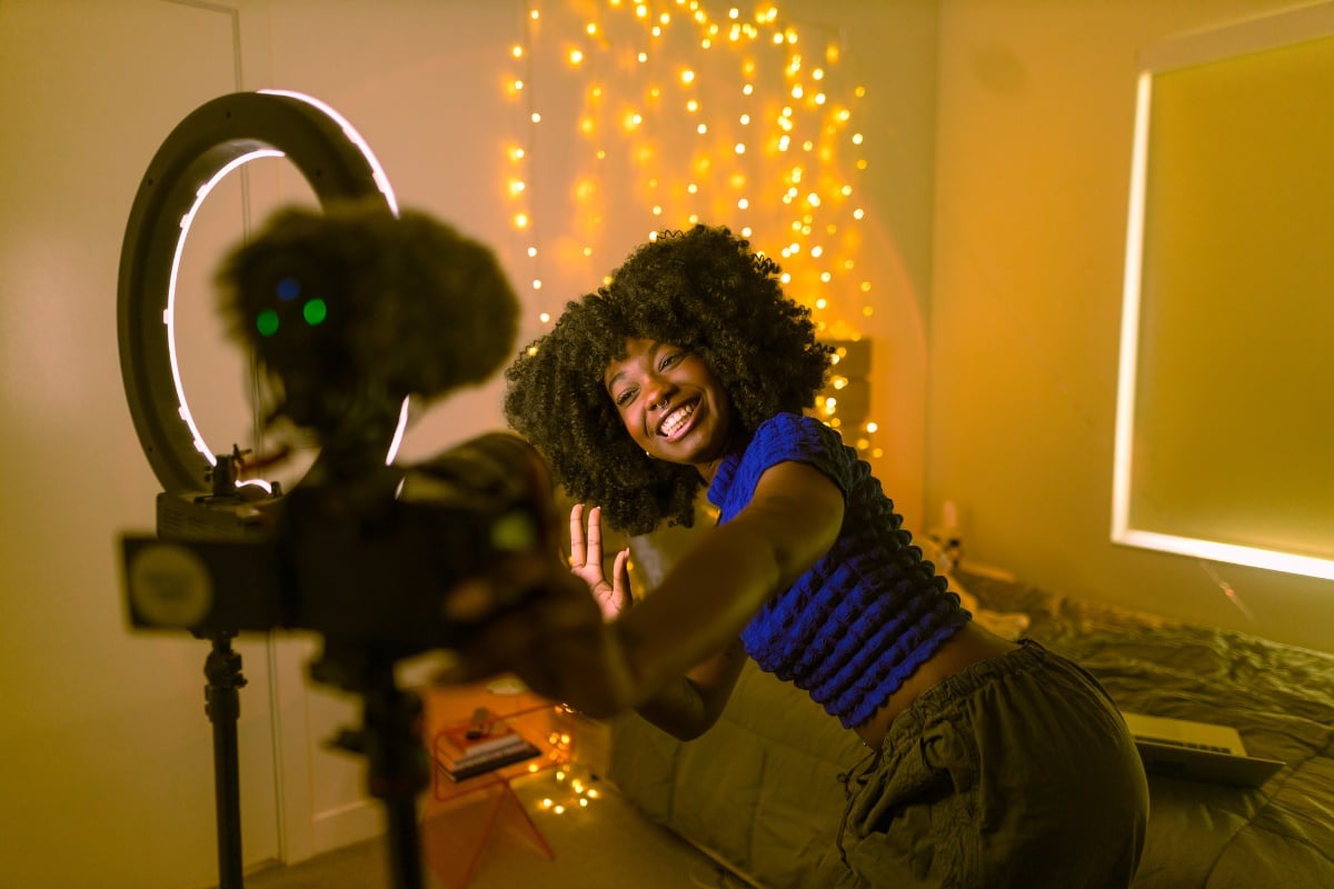 Mulher de pele negra usando cropped azul e calça marrom, em quarto iluminado com luzes na parede, sorrindo em frente a ring light, câmera e microfone