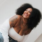 A imagem mostra uma mulher sorridente com cabelo volumoso e crespo. Ela veste blusa branca e jeans, exibe tatuagens nos braços e ombros. A pose é descontraída e alegre, com fundo claro, transmitindo uma sensação de confiança e felicidade.