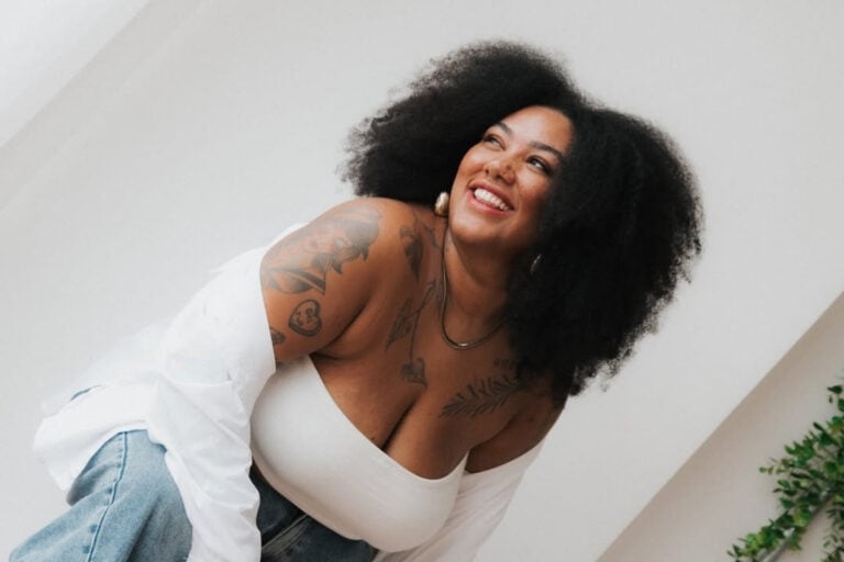 A imagem mostra uma mulher sorridente com cabelo volumoso e crespo. Ela veste blusa branca e jeans, exibe tatuagens nos braços e ombros. A pose é descontraída e alegre, com fundo claro, transmitindo uma sensação de confiança e felicidade.