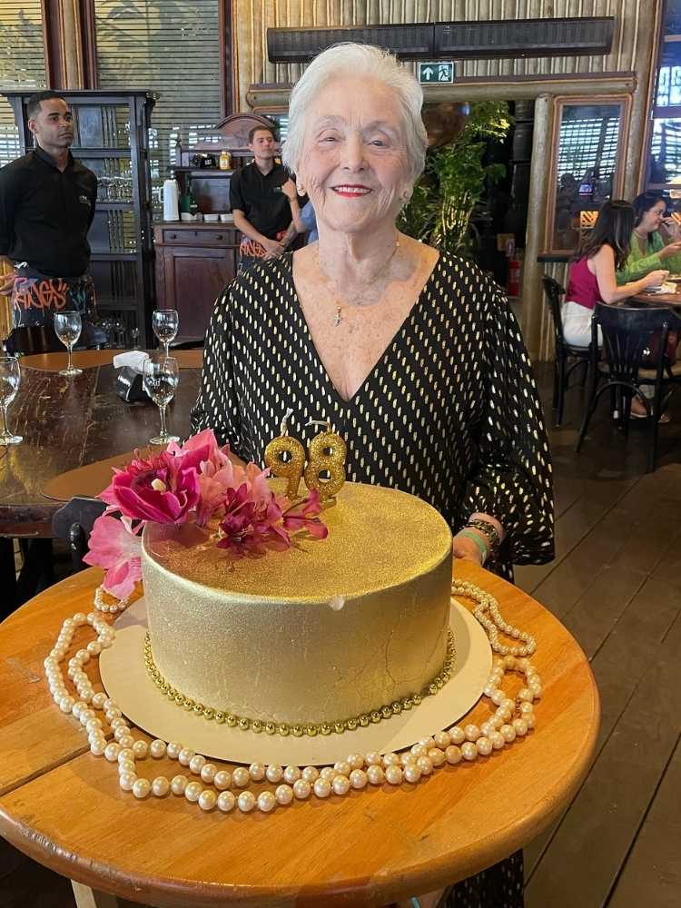 Zuleide Lima usando vestido branco e preto com mesa de madeira com bolo dourado de 98 anos à frente