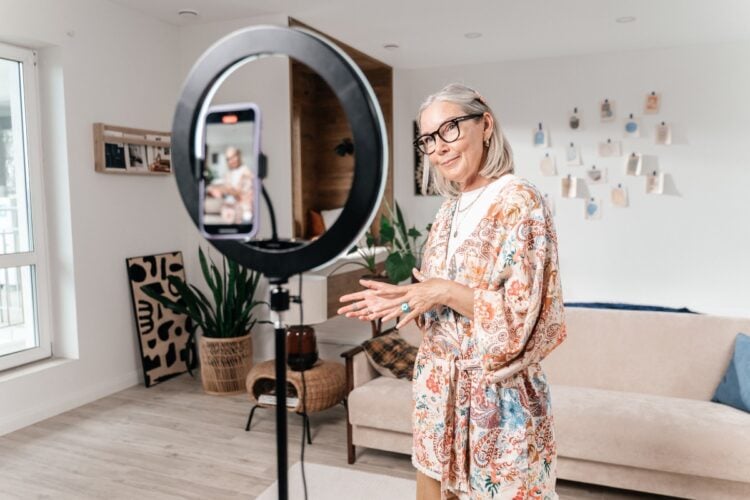 Senhora usando vestido estampado em frente ao celular e ring light, com sala ao fundo, com sofá bege e decorações na parede