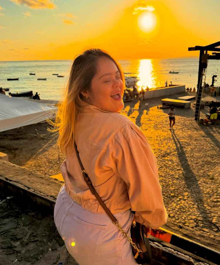 A imagem captura uma mulher sorrindo ao pôr do sol, à beira-mar. Ela veste jaqueta clara e segura uma bolsa. O cenário é vibrante e acolhedor. Representa influenciadores PcD, simbolizando alegria, inclusão e diversidade em um ambiente natural deslumbrante.