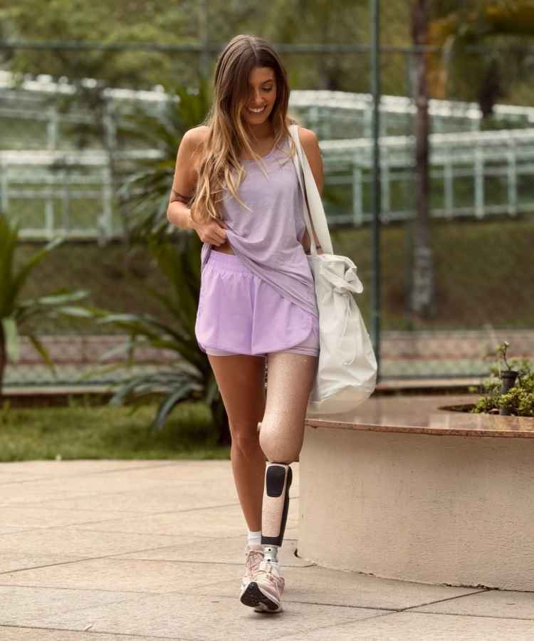 A imagem mostra uma mulher sorridente caminhando com uma prótese na perna. Ela veste roupas esportivas lilás e carrega uma bolsa branca. Ao fundo, há vegetação e uma cerca. Ela representa influenciadores PcD, inspirando inclusão e superação.