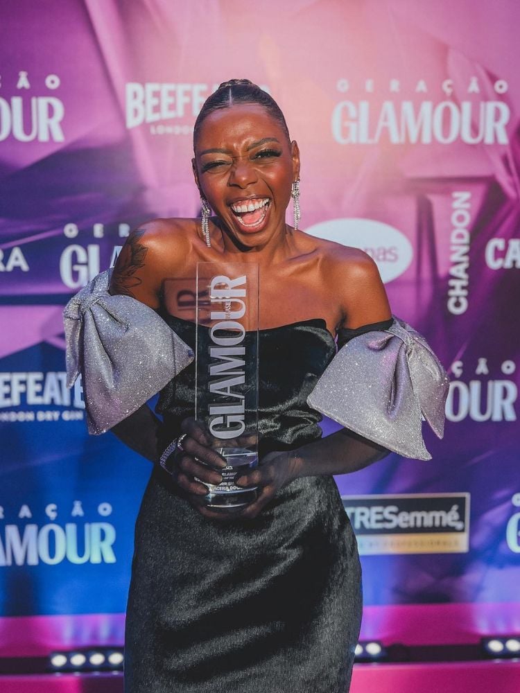 Jacira usando vestido preto sem alças com um laço maxi prateado em cada braço e segurando troféu da Glamour 