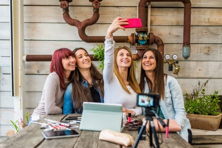 Quatro amigas sorridentes tiram uma selfie com um smartphone rosa. Elas estão sentadas à mesa ao lado de uma câmera, tablet e cosméticos. O ambiente é descontraído, com tubulações antigas e vasos de plantas ao fundo, criando um cenário industrial.