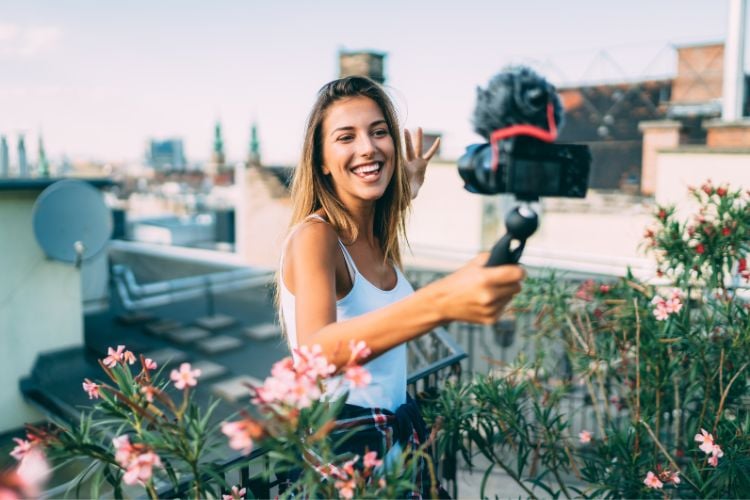 Uma jovem sorri enquanto grava um vídeo com uma câmera em um suporte. Ela está em um terraço rodeado por flores, usando uma regata branca. Ao fundo, há um cenário urbano com antenas e telhados sob um céu claro. marketing de influência