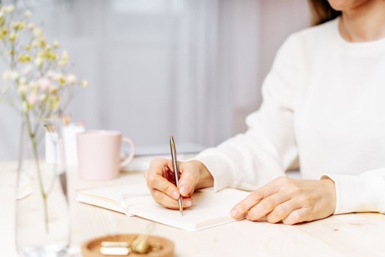 A pessoa, vestindo um suéter branco, escreve em um caderno aberto sobre uma mesa de madeira clara. Ao lado, há uma caneca rosa e um vaso com flores delicadas. O ambiente é iluminado e organizado, transmitindo calma e concentração.