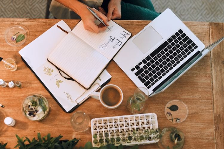 Uma pessoa escreve em um caderno ao lado de um laptop sobre uma mesa de madeira. Há plantas, uma xícara de café e tubos de ensaio. O ambiente é organizado e criativo, sugerindo um espaço de trabalho natural e inspirador.