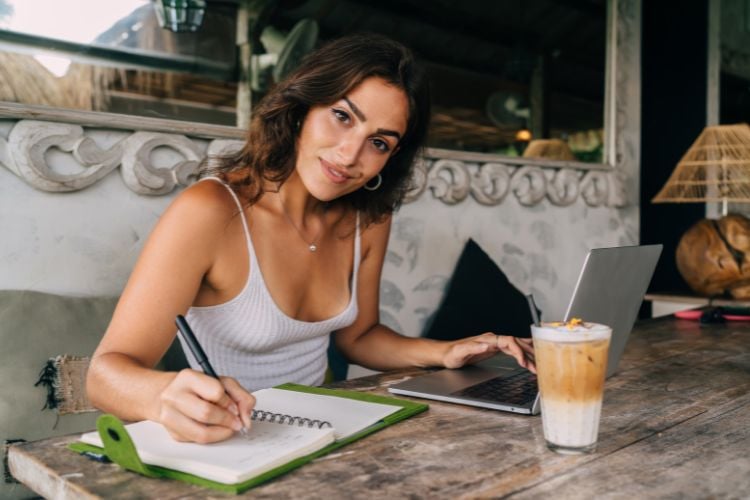 A mulher está sentada em uma mesa de madeira, escrevendo em um caderno com metas para o ano novo. Ela usa uma blusa cinza e olha para a câmera. Ao lado, há um laptop e um copo de café gelado. O ambiente é acolhedor e rústico.