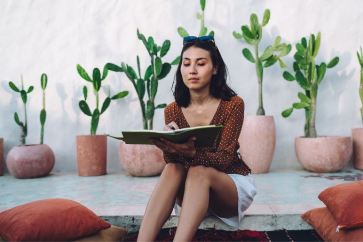 A mulher está sentada, lendo um livro verde com metas para o ano novo. Ela veste uma blusa branca e shorts jeans, em um ambiente ao ar livre com vegetação ao fundo. O clima parece tranquilo, com céu nublado e palmeiras ao redor.