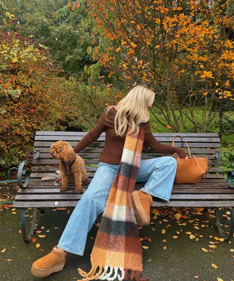 Uma mulher sentada em um banco de parque durante o outono. Ela usa suéter marrom, jeans claros, botas plataforma e cachecol xadrez nas cores outonais. Ao lado, uma bolsa marrom e um cachorro peludo marrom. Folhas laranjas caem ao redor. - look com a cor do anor 2025: Mocha Mousse