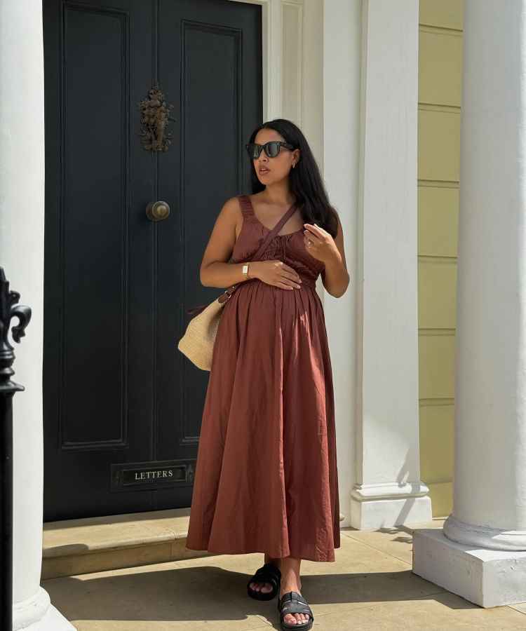 Uma mulher grávida está de pé diante de uma porta preta. Ela usa um vestido longo marrom, sandálias pretas e óculos escuros. Segura uma bolsa de palha e está cercada por colunas brancas, sob luz solar suave criando uma atmosfera tranquila.