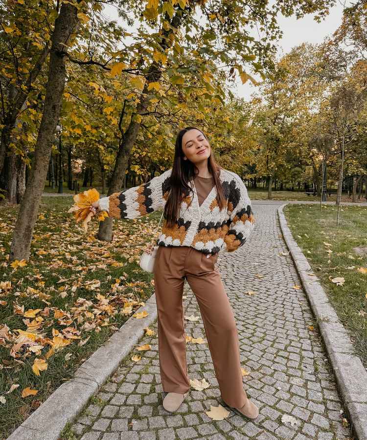 A imagem mostra uma mulher sorridente em um parque durante o outono. Ela veste uma blusa de tricô colorida com desenhos geométricos, calças marrom-terrosas e segura folhas amarelas. O caminho de pedra e as árvores ao fundo criam uma cena acolhedora. - look com a cor do anor 2025: Mocha Mousse