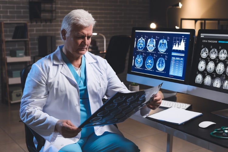 Médico de pele clara usando roupa azul e jaleco, sentado ao lado de mesa com computador, aberto em exames, e segurando raio-x