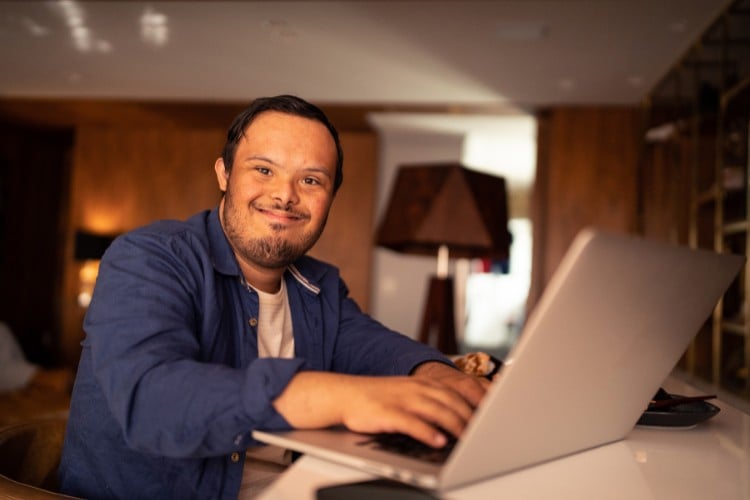 Homem com Síndrome de Down usando blusa branca, camisa azul, sentado à frente de notebook