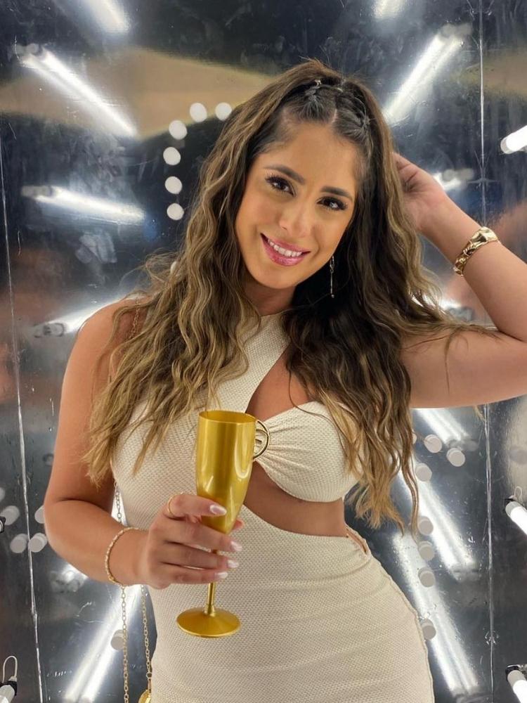 Mulher usando vestido dourado, segurando taça dourada e usando cabelo longo castanho em penteado com triondas