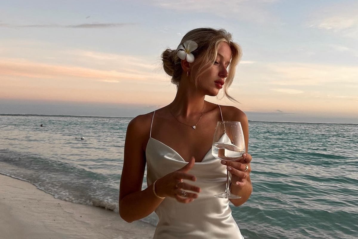 Mulher loira de pele clara usando coque despojado, vestido branco, segurando taça e praia ao fundo