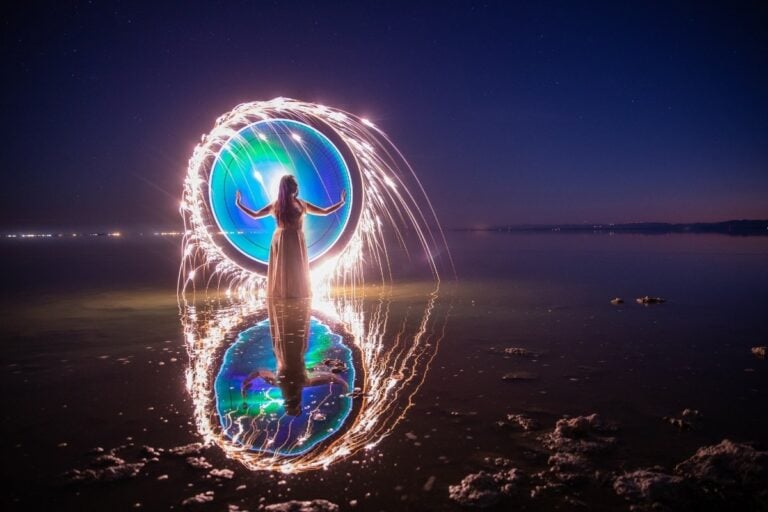 Uma mulher em pé na água, cercada por um círculo luminoso e colorido. O reflexo no lago espelha a cena, criando uma atmosfera mágica e etérea. O céu noturno adiciona um toque de mistério à paisagem.