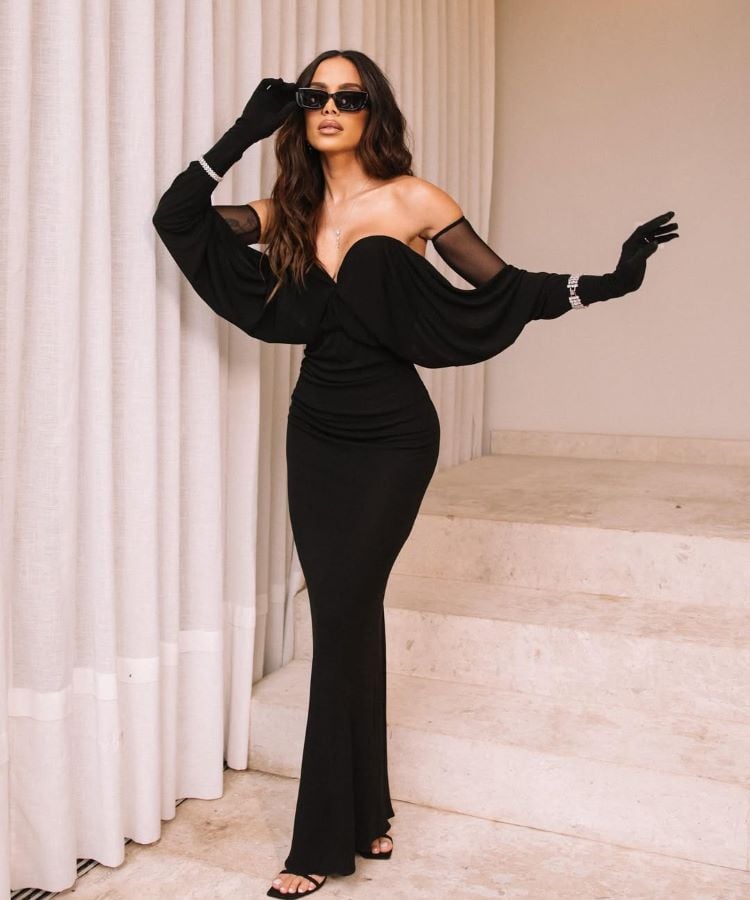 Anitta elegante usa vestido preto longo e justo com luvas combinando para o Prêmio Multishow 2024. Ela está em uma escada, usando óculos escuros e posando confiante ao lado de cortinas brancas.