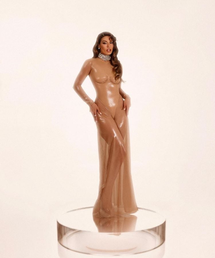 Beatriz Reis em vestido nude brilhante, longo, ajustado e transparente, em um pedestal, para o Prêmio Multishow 2024 looks. Usa colar prateado grande. Cabelo solto, ondulado. Fundo claro e iluminação suave. Pose confiante e elegante.