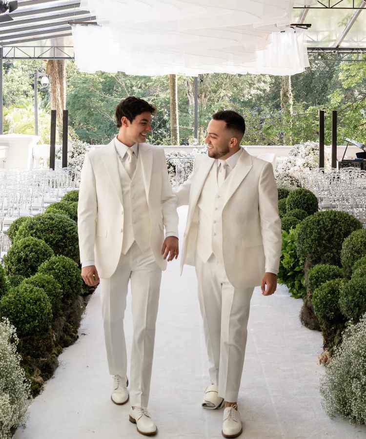 Lucas Rangel e Bley caminham sorridentes, usando ternos brancos combinados, em um ambiente decorado para casamento. Cercados por folhagens verdes e flores brancas, eles trocam olhares de felicidade em um corredor elegante, sob um teto de tecido delicado e translúcido.