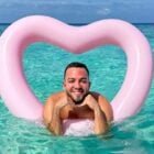 Lucas Rangel sorridente posa em águas cristalinas, apoiado em uma boia rosa em formato de coração. O céu azul complementa o cenário tropical e relaxante, transmitindo uma sensação de alegria e tranquilidade em um ambiente paradisíaco.