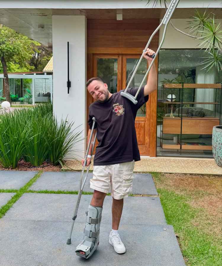 Lucas Rangel sorri enquanto posa em frente a uma casa moderna, usando muletas e uma bota ortopédica na perna esquerda. Vestindo uma camiseta preta e bermuda branca, ele exibe uma atitude positiva em um ambiente verde e acolhedor.