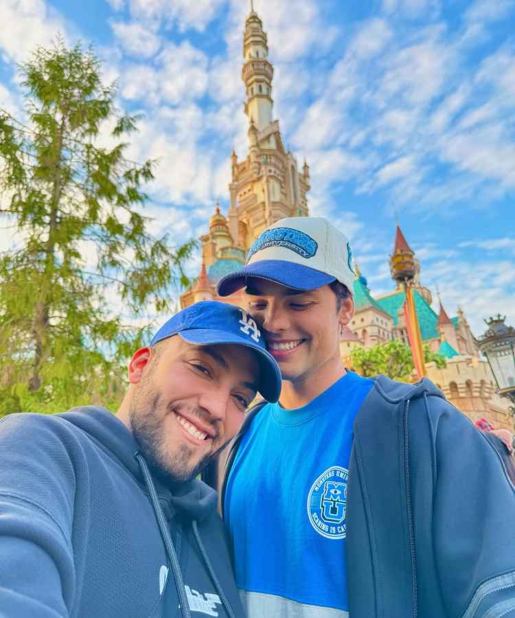 Lucas Rangel e Bley sorridentes posam juntos em frente a um castelo em um parque temático. Ambos usam bonés e roupas casuais, enquanto o céu azul repleto de nuvens adiciona um toque mágico à cena, capturando um momento de alegria e união.