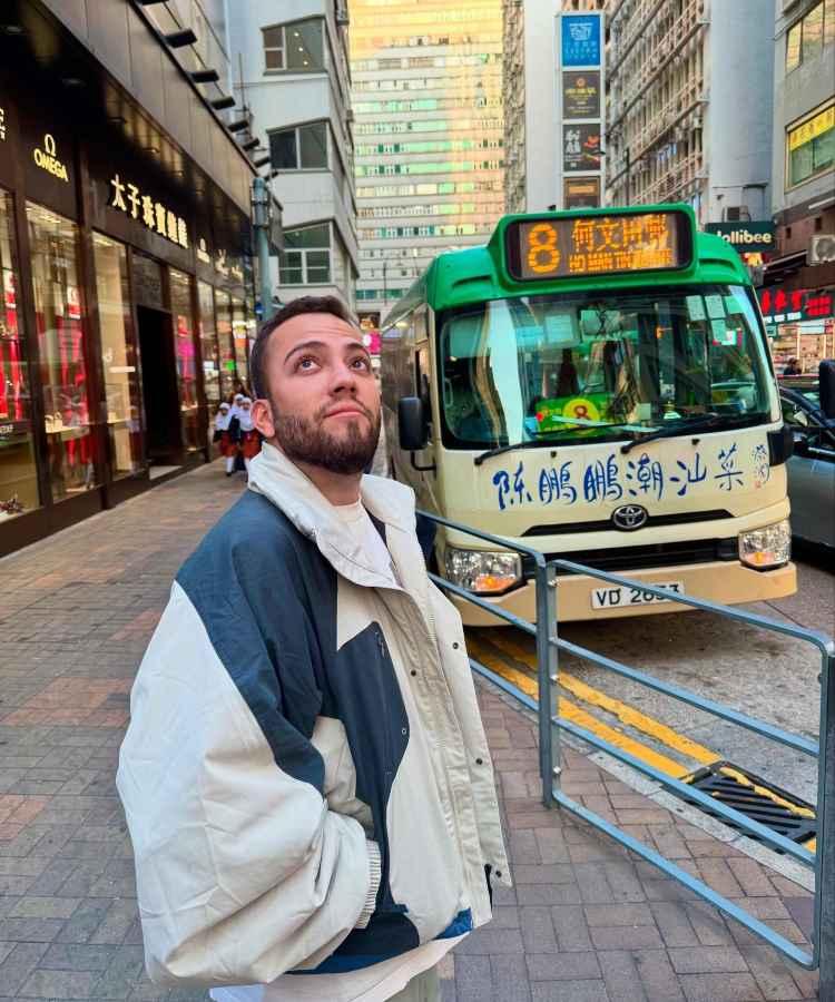 Um homem olha para cima em uma rua movimentada de uma cidade asiática. Ele veste uma jaqueta oversized e está próximo de um micro-ônibus verde com caracteres chineses. Ao fundo, lojas e prédios altos completam a cena urbana vibrante.