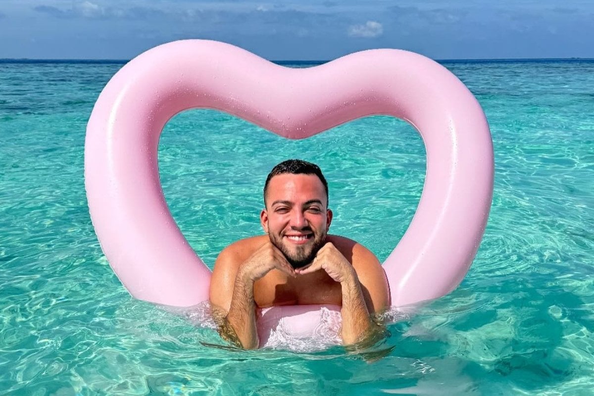 Lucas Rangel sorridente posa em águas cristalinas, apoiado em uma boia rosa em formato de coração. O céu azul complementa o cenário tropical e relaxante, transmitindo uma sensação de alegria e tranquilidade em um ambiente paradisíaco.