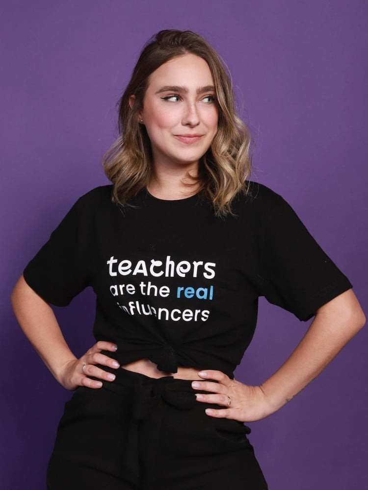 Teacher Paula Gabriela usando blusa preta escrito "teachers are the real influencers" em fundo roxo 