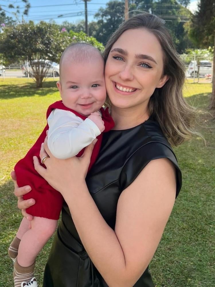 Paula Gabriela usando blusa preta de couro e segurando bebê que usa roupas brancas e vermelhas