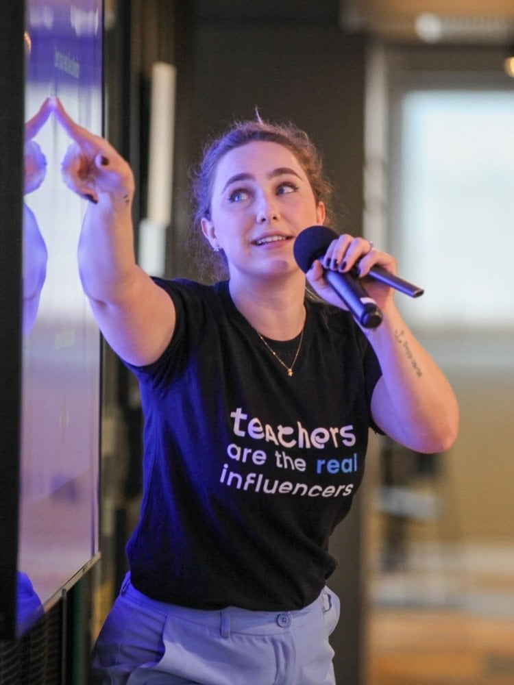 Teacher Paula usando camiseta preta escrito "teachers are the real influencers" enquanto aponta para algo no telão e segura microfone