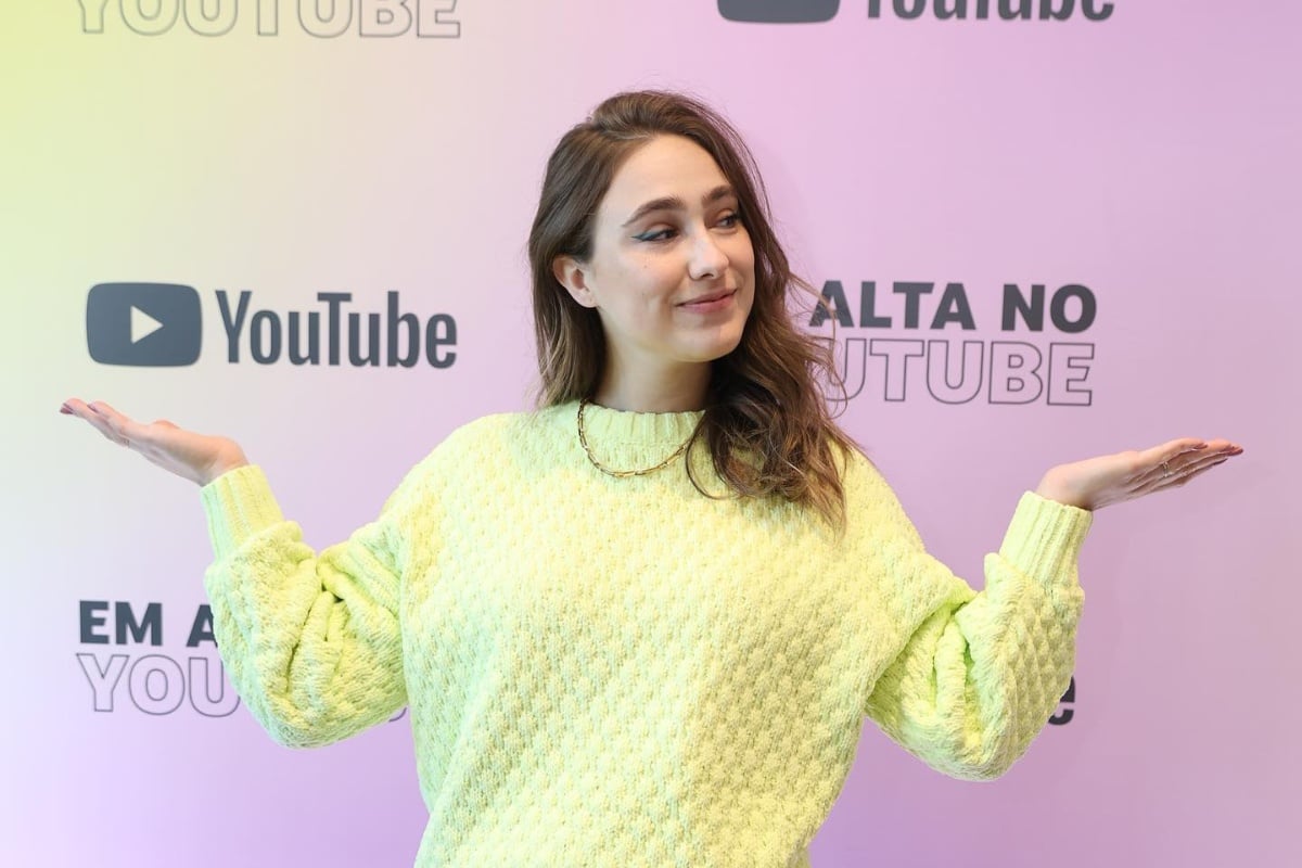 Teacher Paula Gabriela usando suéter amarelo em frente a painel do YouTube