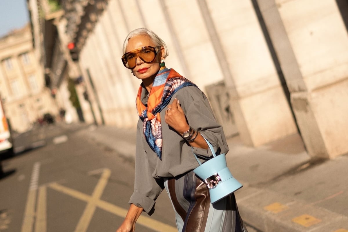 Senhora de pele clara usando óculos aviador, lenço colorido, blusa cinza, acessórios em prata e dourado e bolsa azul