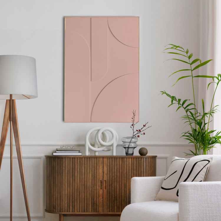 A imagem mostra uma sala elegante com parede branca e arte abstrata em rosa pálido. Há um aparador de madeira com decoração minimalista, incluindo vasos e livros. Uma luminária de tripé e plantas verdes adicionam sofisticação ao ambiente acolhedor.  - tendências de decoração para 2025