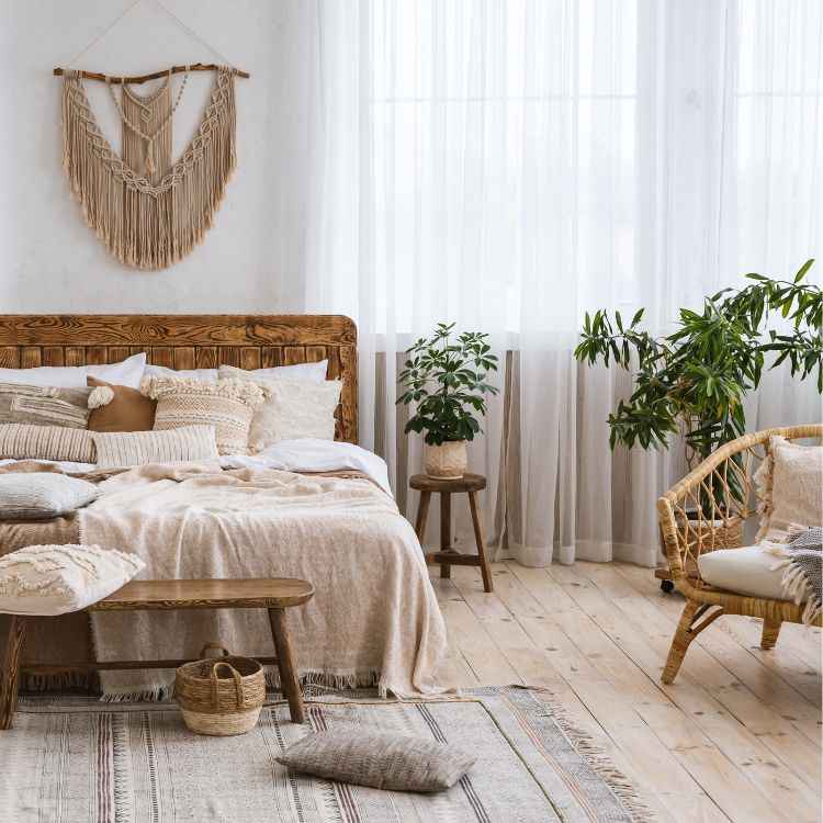 O quarto exibe um estilo boho com cama de madeira adornada por almofadas e colchas em tons neutros. Nas paredes, uma peça de macramê complementa a decoração. Plantas verdes e uma cadeira de vime acrescentam um toque natural ao ambiente iluminado.