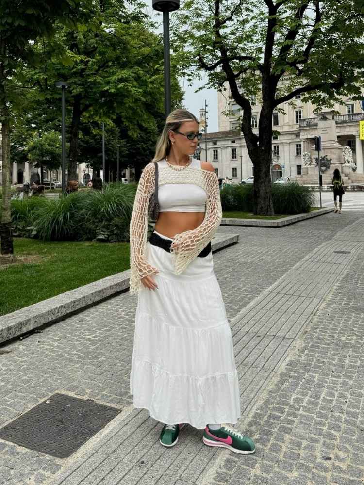 Mulher loira de pele clara em praça usando cropped branco, saia longa branca, cinto preto e Nike Cortez verde e rosa