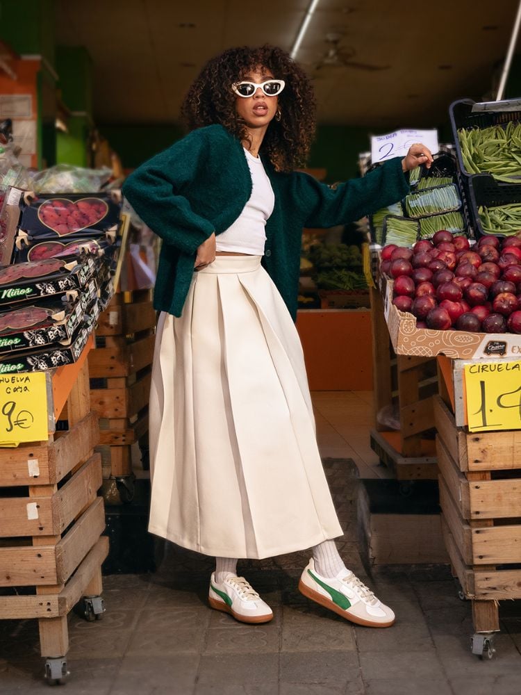 Mulher de pele negra em feira usando cropped branco, casaco verde, saia midi bege e Puma Palermo branco e verde