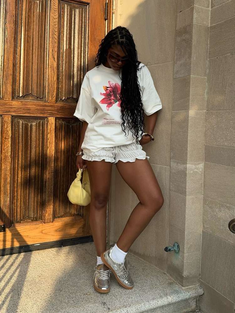 Mulher de pele negra usando camiseta branca com flor rosa, shorts branco franzido, bolsa amarela e Steve Madden Madrid prateado