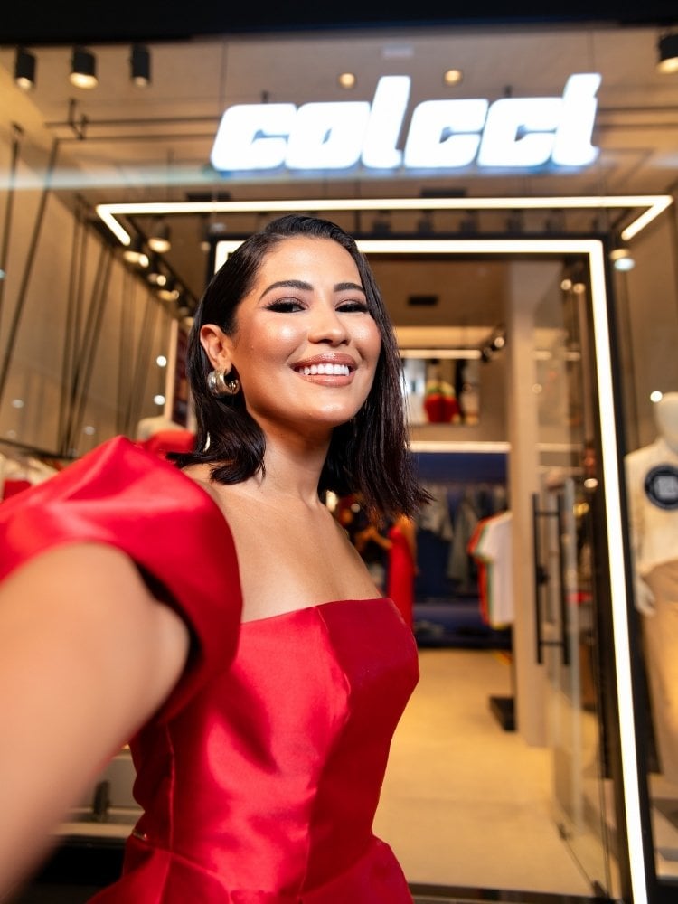 Thaynara OG sorridente em vestido vermelho faz selfie em frente à entrada da loja Colcci. Cabelo escuro, maquiagem elegante. Interior da loja iluminado, moderno. Logotipo destacado acima. Atmosfera de moda e sofisticação.