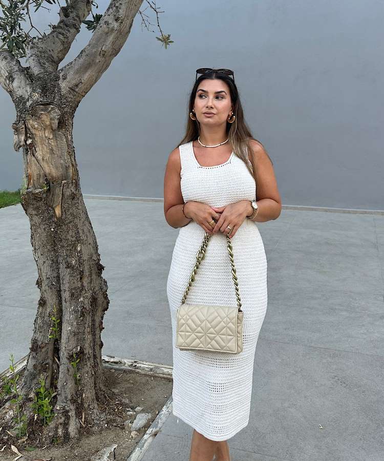 Mulher com vestido de crochê branco, bolsa, ao lado de árvore.