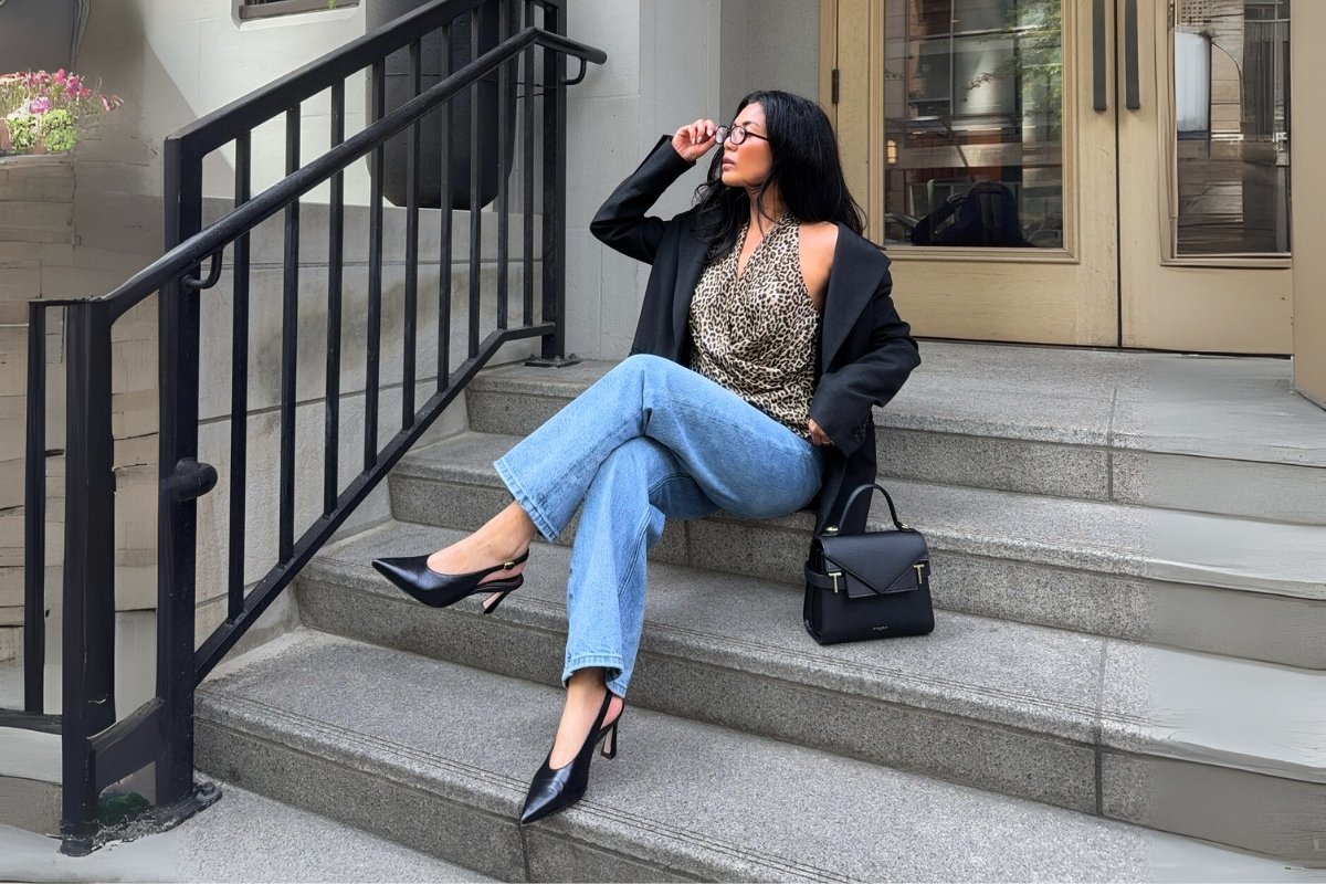 Mulher com óculos, blazer preto, blusa estampada de onça e calça jeans, sentada em escadas de pedra. Ela usa sapatos pretos de salto agulha e segura uma bolsa preta. A imagem é de corpo inteiro.